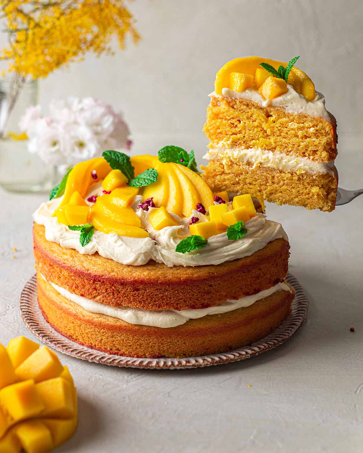 Vegan mango cake topped with cream cheese frosting and fresh mango. Slice of cake is lifted up showing fluffy texture.