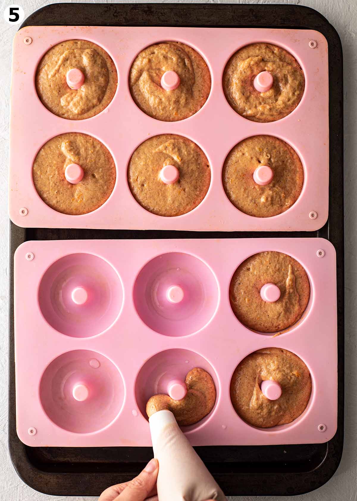Two silicone donut moulds on tray with hands piping thick pumpkin donut batter into each donut shape.
