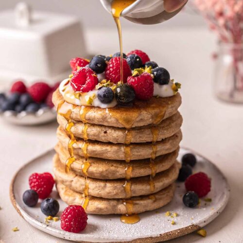 Tall stack of decorated whole wheat pancakes with maple syrup poured on top and dripping down sides.