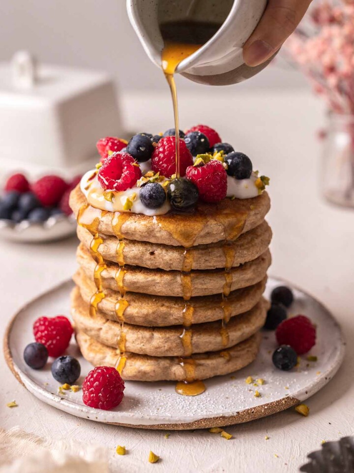 Tall stack of decorated whole wheat pancakes with maple syrup poured on top and dripping down sides.