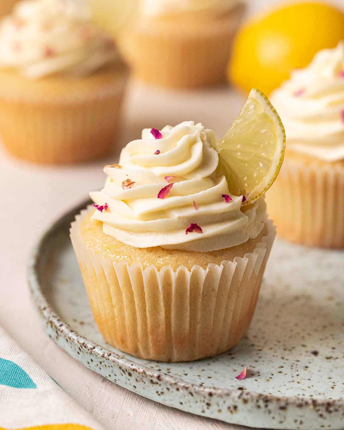 Close up of vegan lemon cupcake with tall frosting swirl and is decorated with lemon segment and rose petals.