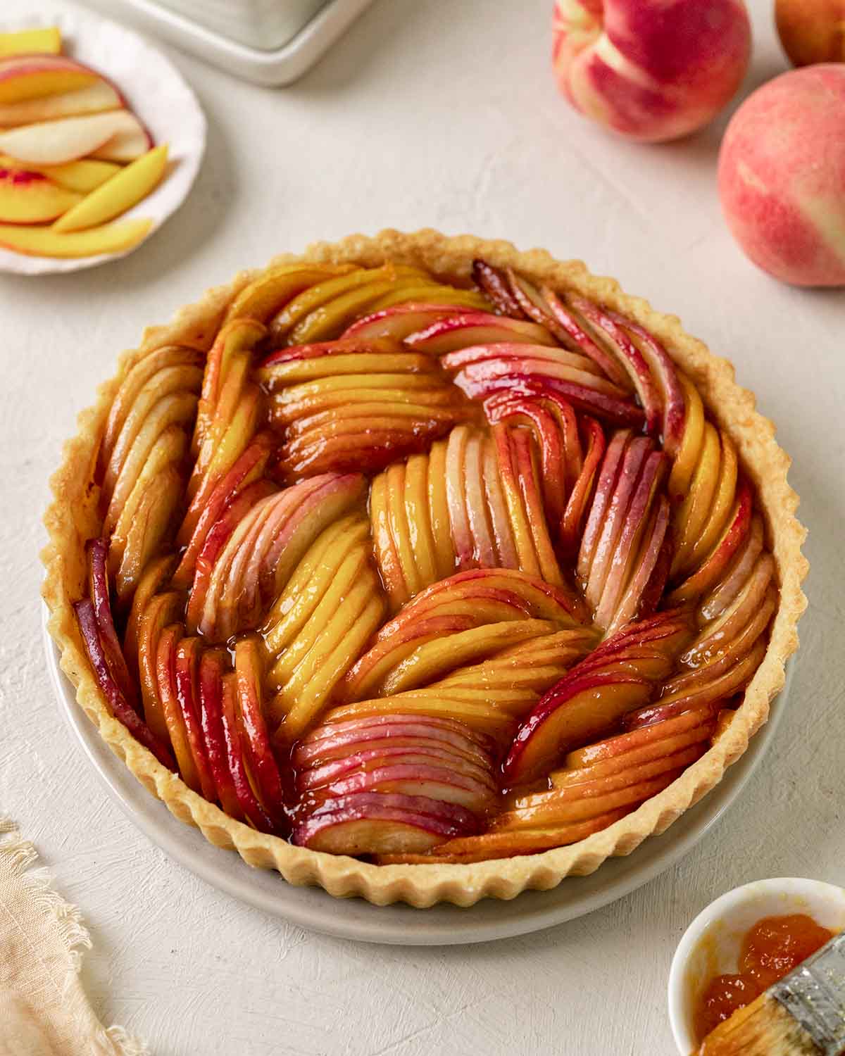 Baked tart consisting of thinly sliced peaches in a buttery pastry shell. Peaches have a thick sticky glaze on top.