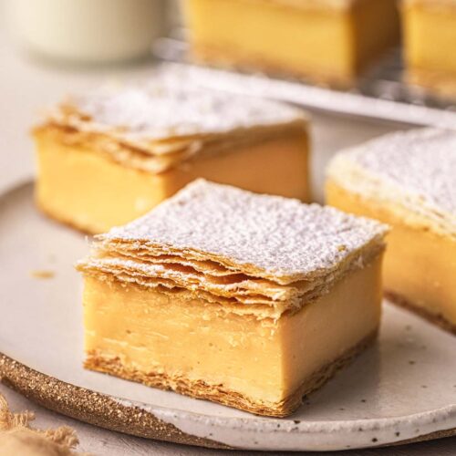 Close up of vanilla slice on a plate. The slice has two flaky layers of puff pastry sandwiching a thick layer of custard.