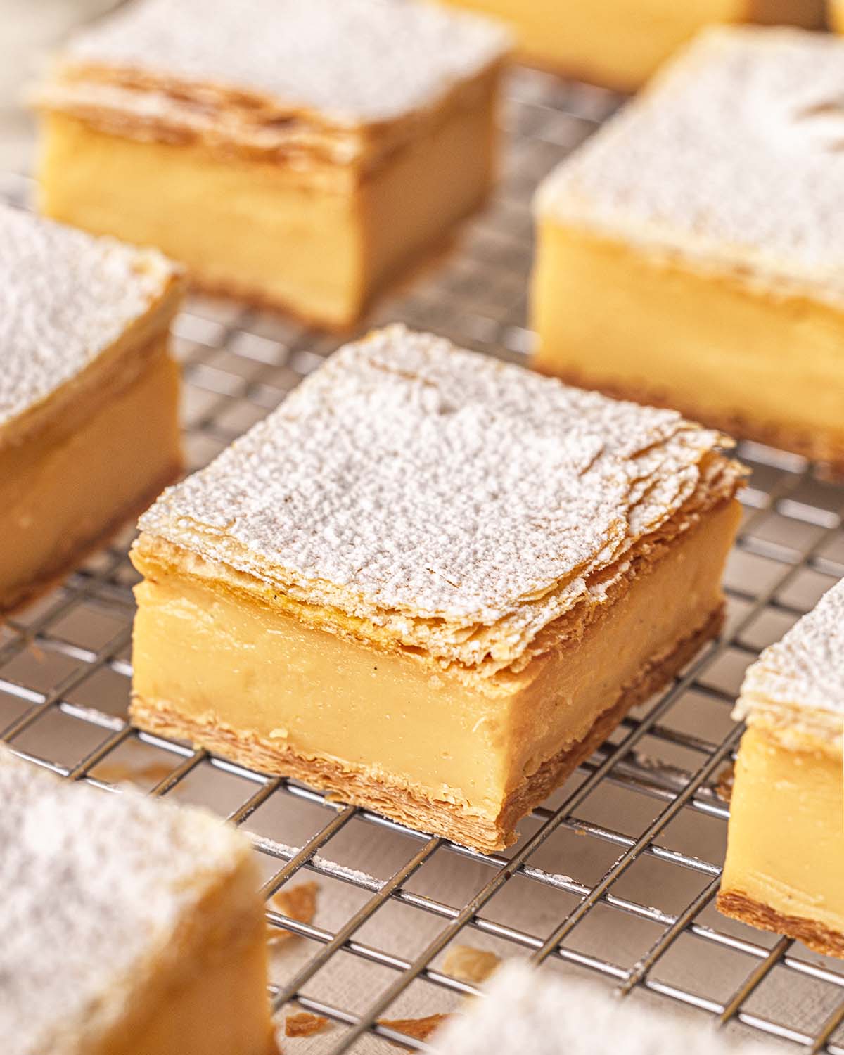 Close up of rows of vanilla slices on wire rack. Each slice is is genorously dusted with icing sugar.