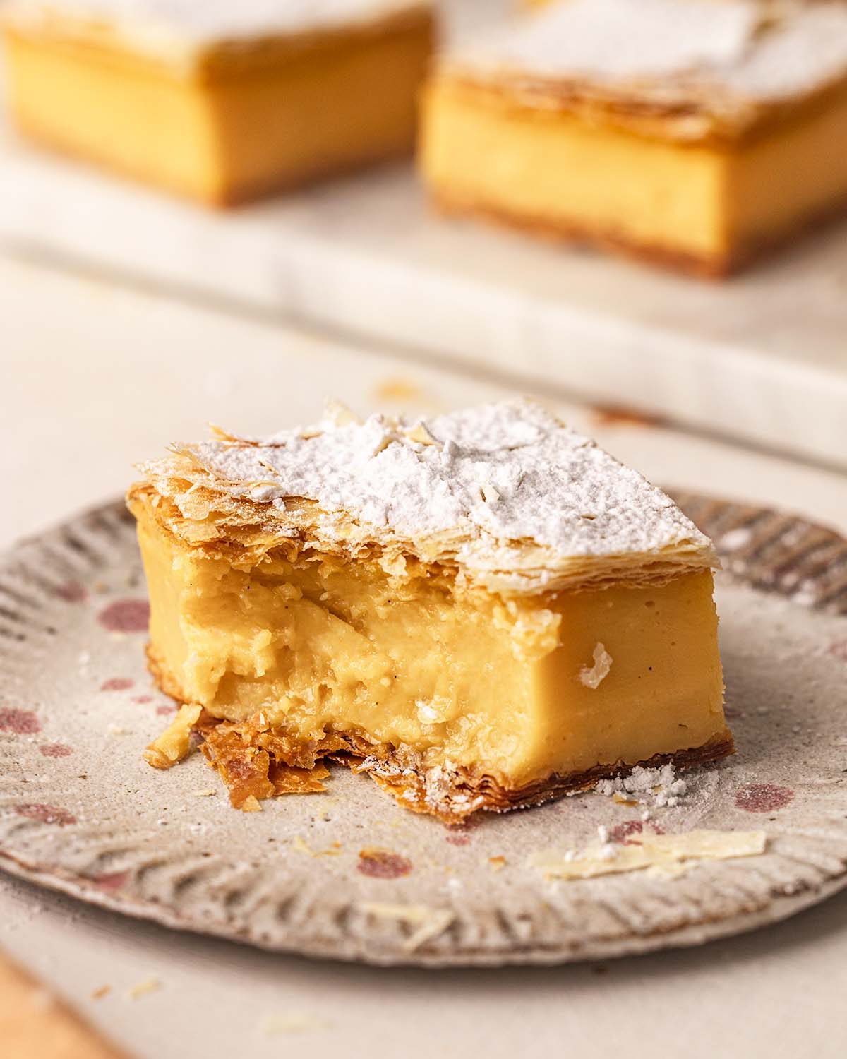 Close up of vanilla slice on plate with bite taken out showing creamy custard and layers of flaky pastry.