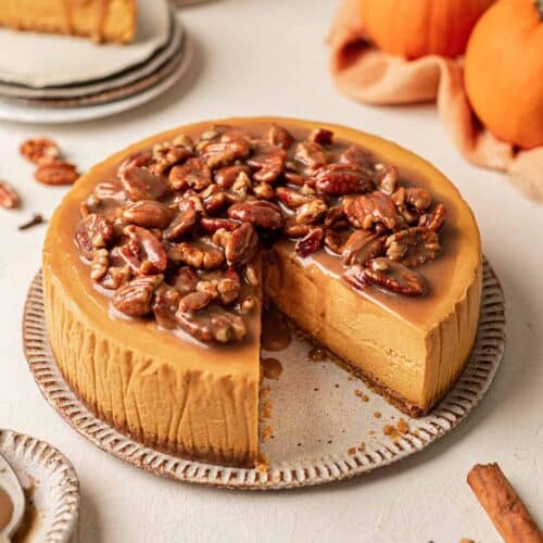 Vegan pumpkin cheesecake topped with a pecan caramel sauce. The cheesecake is on plate and there's a slice taken out to reveal the smooth creamy texture of the cheesecake.