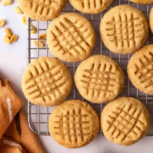 Peanut butter cookies in single layer on wire rack with peanuts, peanut butter and brown tea towel on the side.
