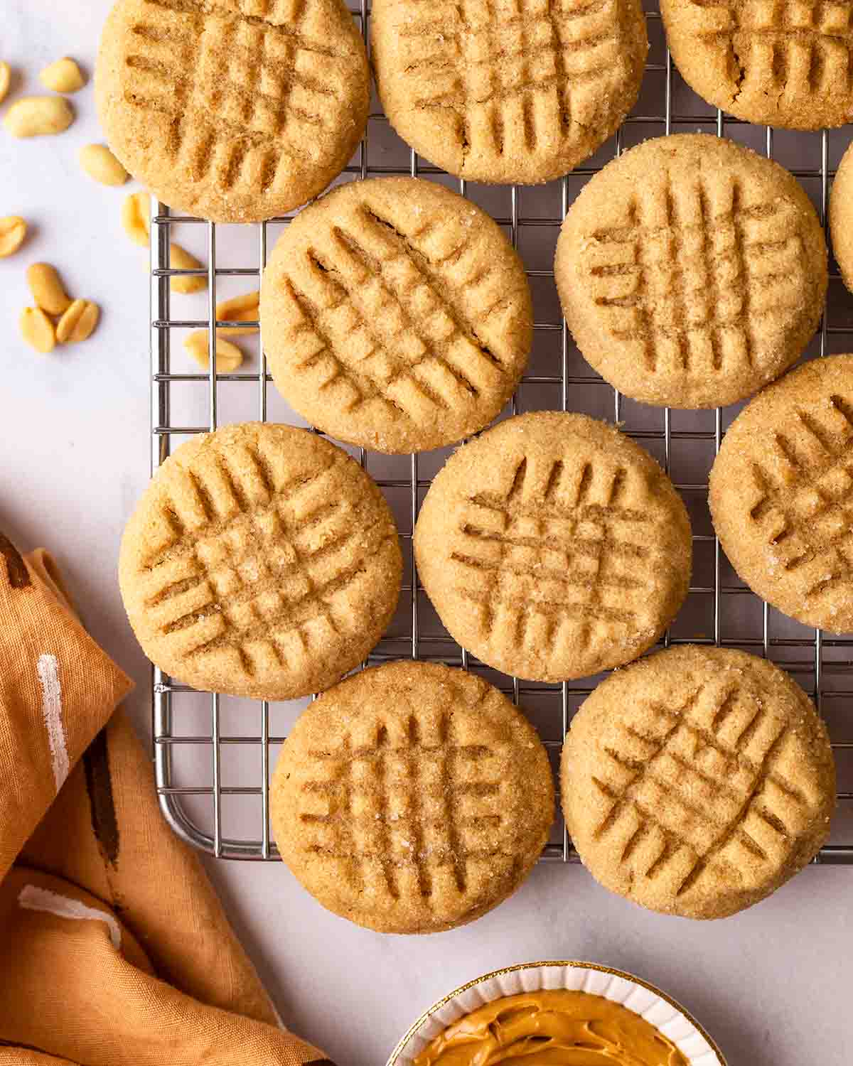 Peanut butter cookies in single layer on wire rack with peanuts, peanut butter and brown tea towel on the side.