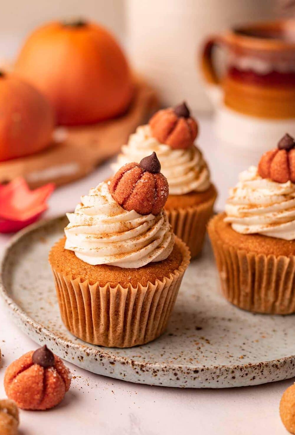 Three pumpkin cupcakes on plate surrounded by mini pumpkins. The cupcakes are decorated with frosting and edible pumpkin decorations.