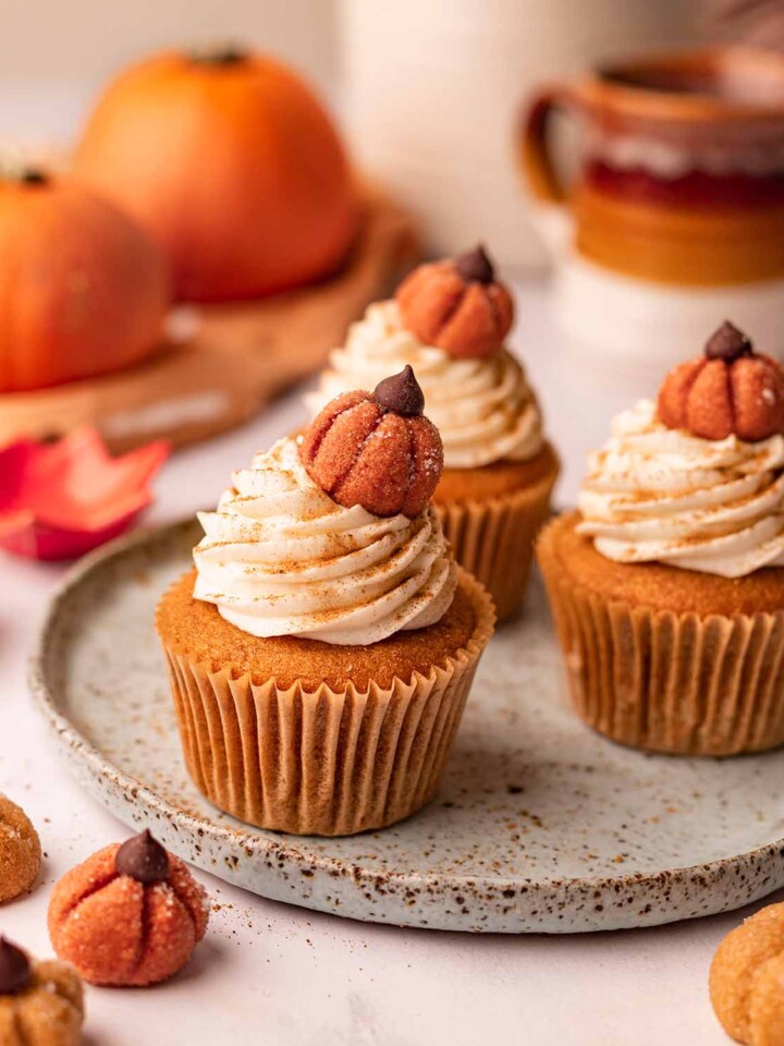Three pumpkin cupcakes on plate surrounded by mini pumpkins. The cupcakes are decorated with frosting and edible pumpkin decorations.