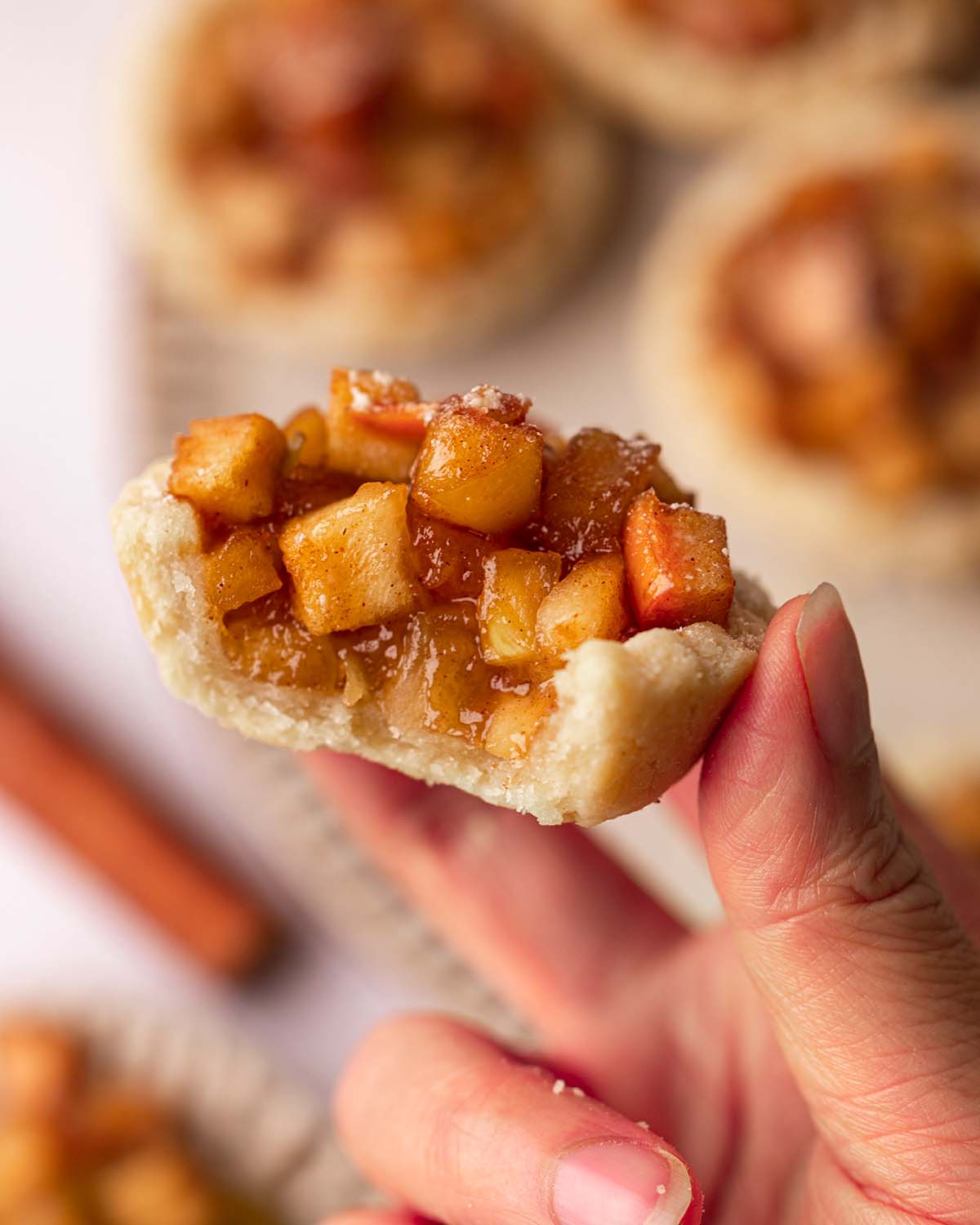 Hand holding a mini apple tart that has been bitten into. Image focuses on gooey apple filling of the tart.