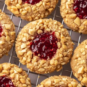 Close up of peanut butter jelly cookies on cooling rack. Each cookie is coated in chopped peanuts and has a thumbprint filled with red jam.