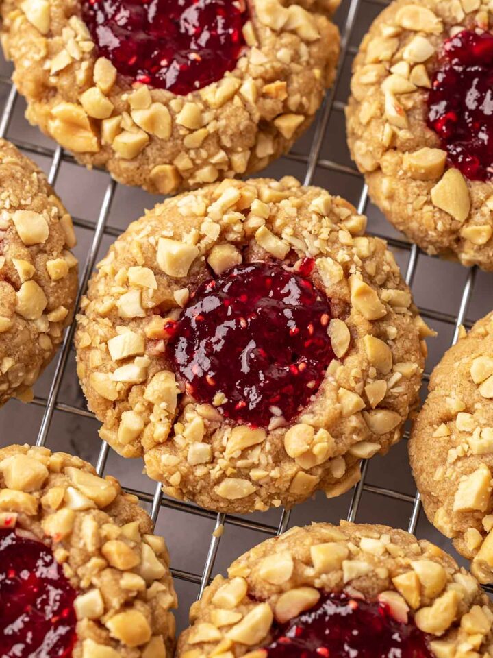 Close up of peanut butter jelly cookies on cooling rack. Each cookie is coated in chopped peanuts and has a thumbprint filled with red jam.