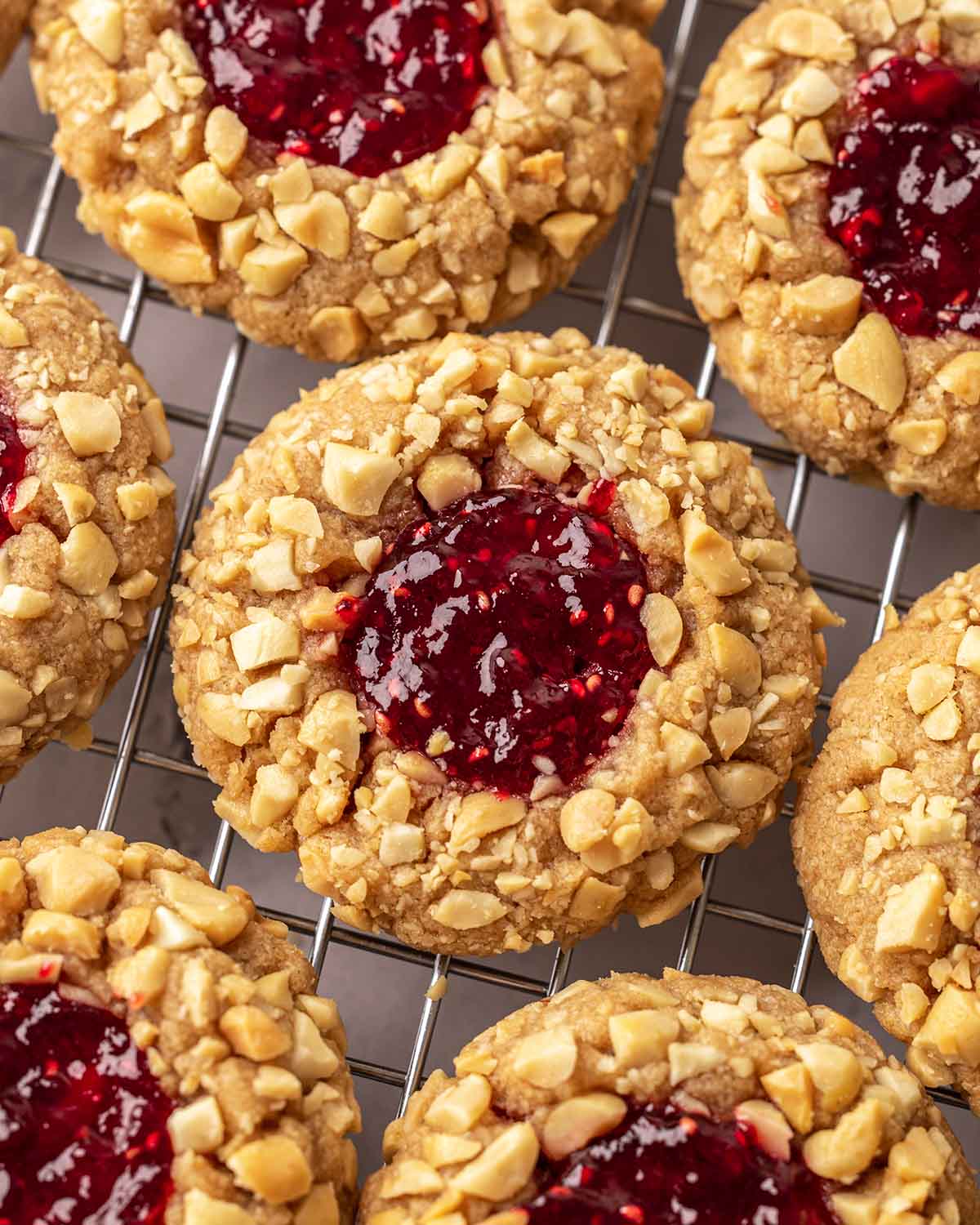Close up of peanut butter jelly cookies on cooling rack. Each cookie is coated in chopped peanuts and has a thumbprint filled with red jam.