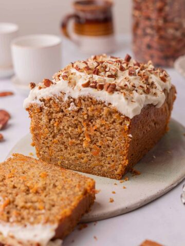 Carrot cake loaf on serving board decorated with white frosting and chopped pecans. Loaf has slice cut out revealing the fluffy coarse texture.