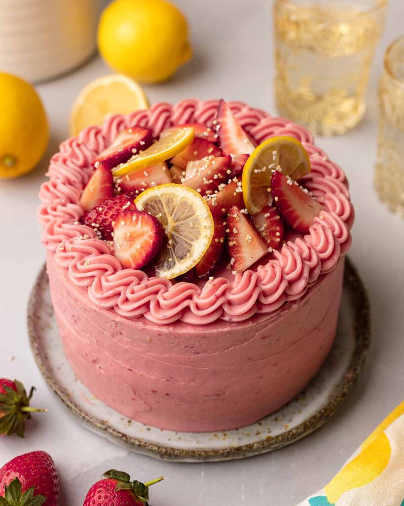 Whole decorated cake on plate. Cake has pink frosting and is topped with fresh strawberries and lemon slices.