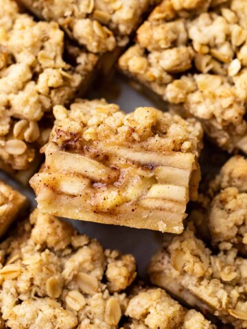Apple pie bars cut into squares. One square is on its side showing the gooey layers, shortcrust pastry and crumble topping.