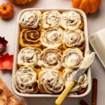 Golden pumpkin cinnamon rolls in baking tray. All rolls except one are covered in cream cheese frosting except one.