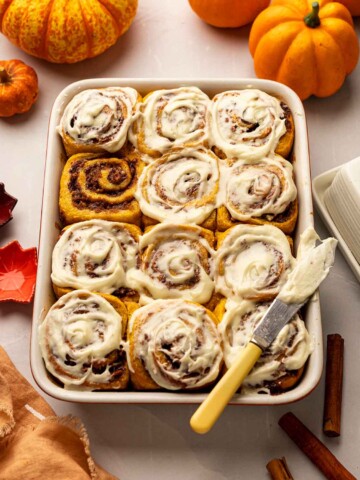 Golden pumpkin cinnamon rolls in baking tray. All rolls except one are covered in cream cheese frosting except one.