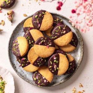 Almond flour shortbread stacked on a plate. The cookies are half dipped in chocolate with a sprinkle of pistachios and dried rose petals.