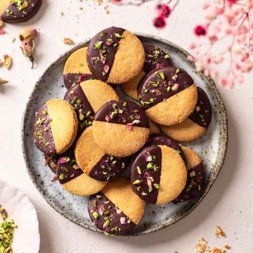 Almond flour shortbread stacked on a plate. The cookies are half dipped in chocolate with a sprinkle of pistachios and dried rose petals.