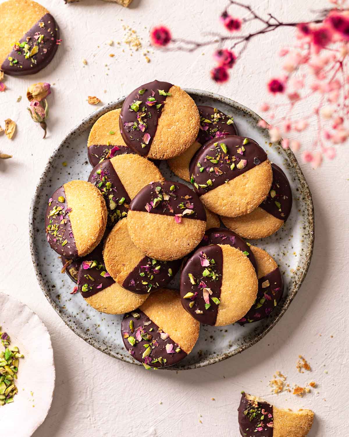 Almond flour shortbread stacked on a plate. The cookies are half dipped in chocolate with a sprinkle of pistachios and dried rose petals.