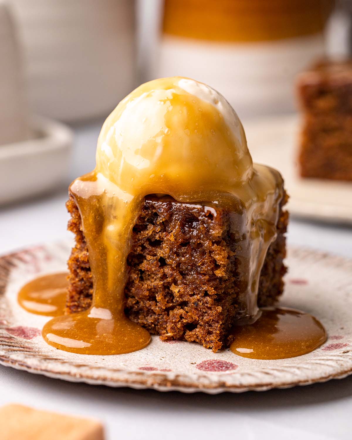Square of sticky date pudding with scoop of ice cream with toffee sauce poured on top.