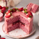 Small strawberry cake on plate with slices cut out revealing the cake layers and jammy filling.