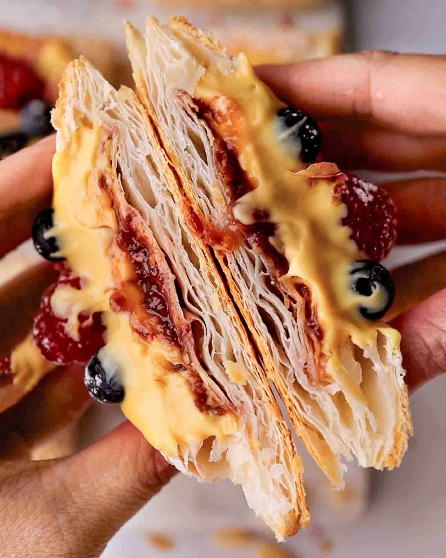 Hands holding two halves of a custard pastry cut showing very flaky layers and creamy custard.