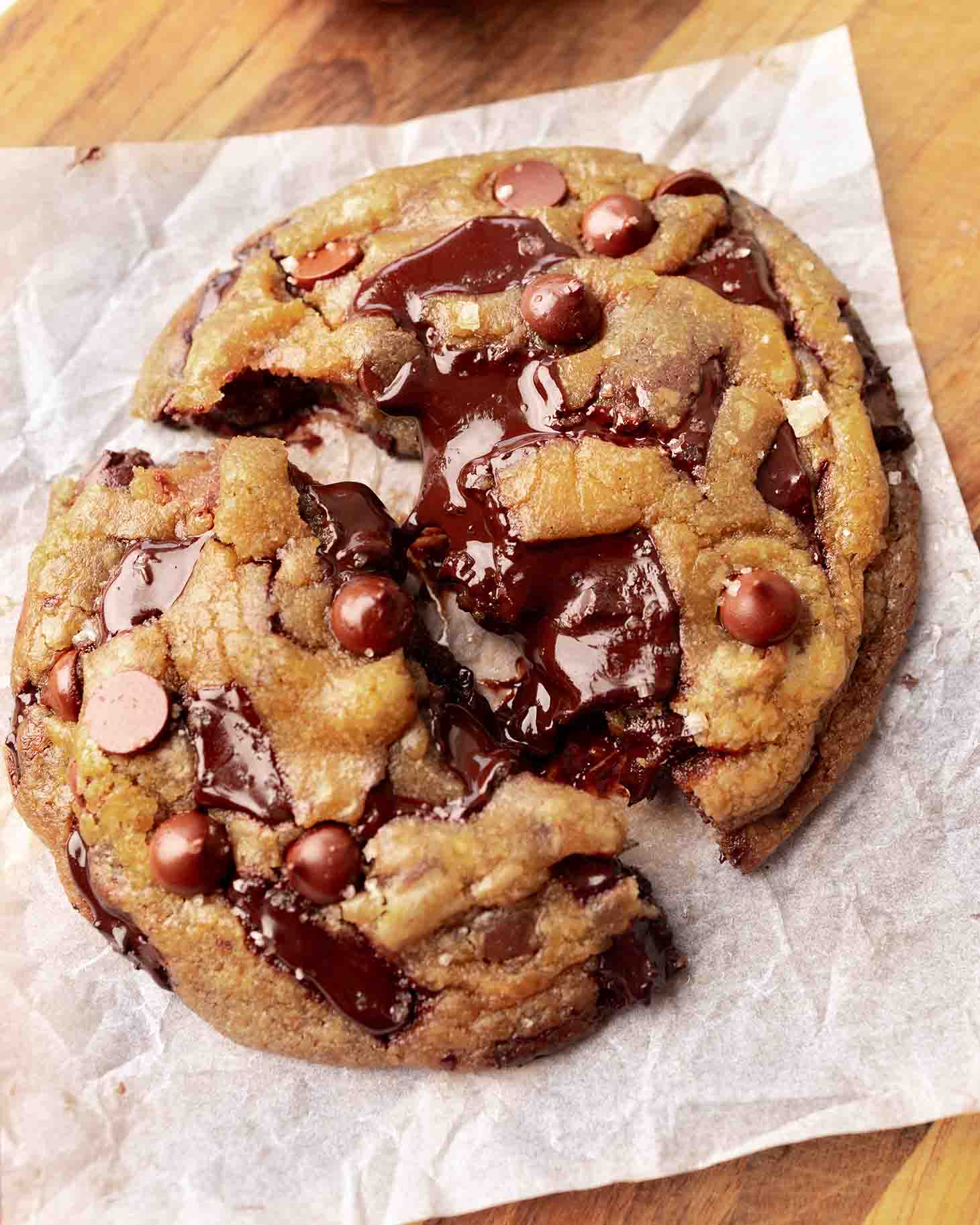 Large vegan cookie broken in half on parchment paper with melty pools of chocolate.